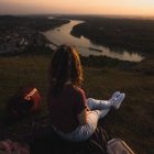Woman sits on hillside overlooking river and town at sunset, creating a serene landscape scene.