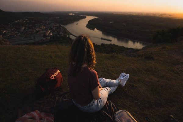 Woman sits on hillside overlooking river and town at sunset, creating a serene landscape scene.