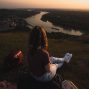 Woman sits on hillside overlooking river and town at sunset, creating a serene landscape scene.